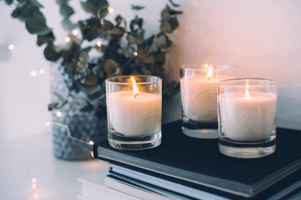 Fotografia de três velas brancas acesas, dentro de recipientes de vidro transparente, posicionadas sobre uma pilha de livros de capas escuras. Ao fundo, há um vaso com folhas verdes e pequenas luzes decorativas desfocadas. As chamas iluminam suavemente o ambiente, que tem fundo claro e aparência aconchegante.
