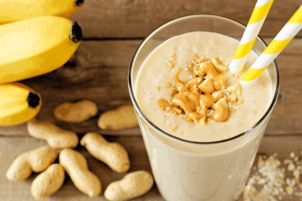 A imagem mostra uma vitamina de banana com amendoim, servida em um copo de vidro sobre uma mesa de madeira. A bebida apresenta textura cremosa e tonalidade clara, sendo finalizada com pedaços de amendoim, aveia e uma porção de pasta de amendoim. Dois canudos listrados em amarelo e branco complementam a apresentação. Ao redor do copo, aparecem bananas maduras, amendoins e flocos de aveia, destacando alguns dos ingredientes usados no preparo.