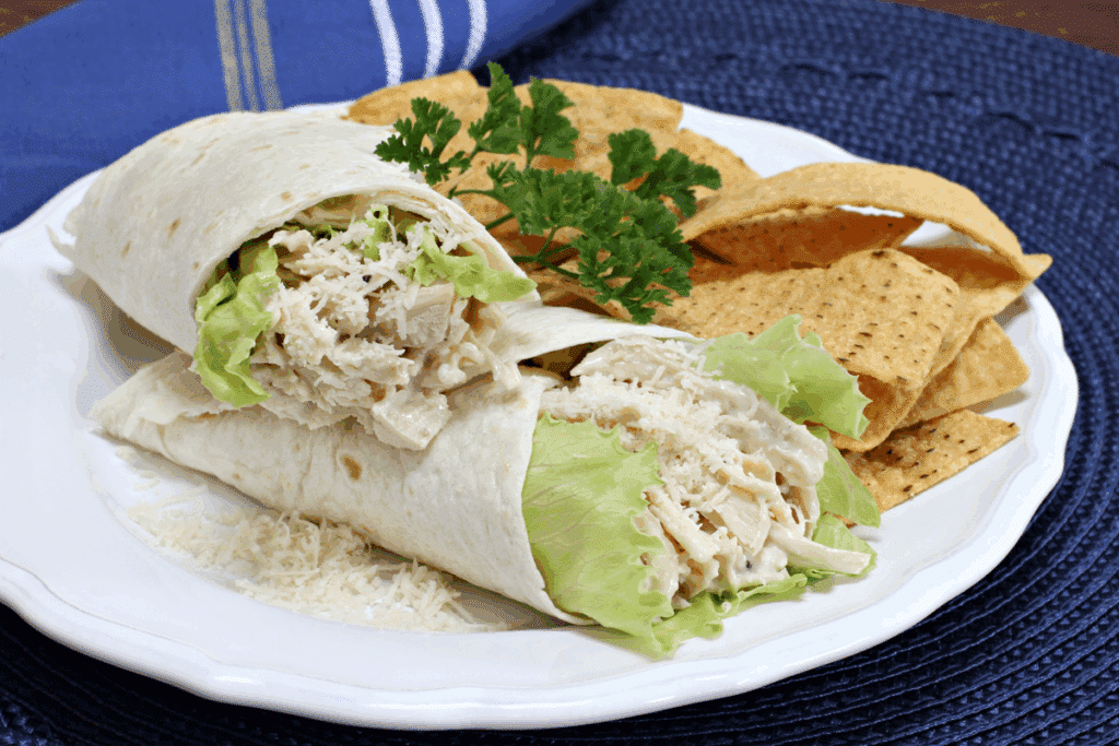 Dois wraps de frango desfiado cortados ao meio, com alface e queijo parmesão ralado, acompanhados de chips de tortilha em um prato branco.